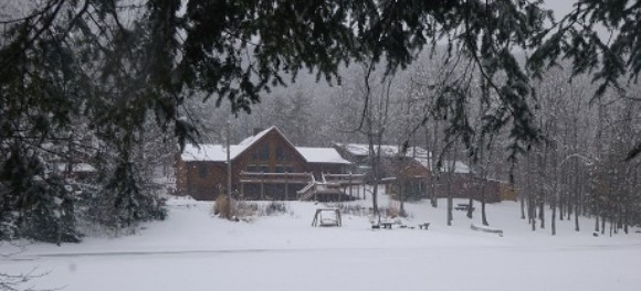 A wintry scene at Pioneer Mountain Homestead. The homestead's main house is seen from across the pond. A blanket of snow has covered everything.